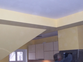 Wnętrze w trakcie prac wykończeniowych w Poznaniu: żółte ściany z szarym pasem dekoracyjnym pod sufitem, widoczne szafki kuchenne i fragment osoby pracującej.